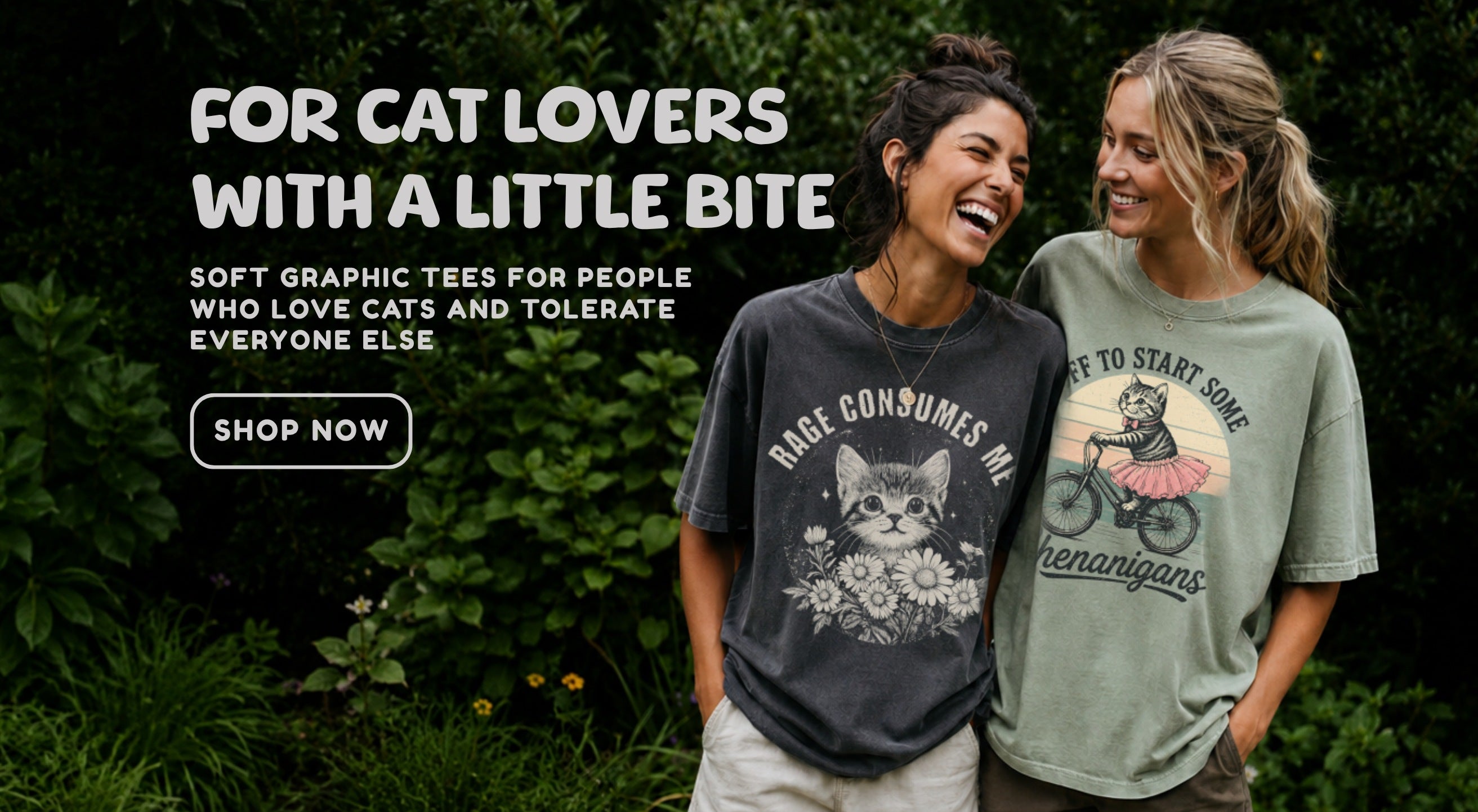 Hero banner for cat lover graphic tees showing two smiling women in oversized cat-themed t-shirts standing in a lush green garden, with text reading “For cat lovers with a little bite” and a Shop Now button.